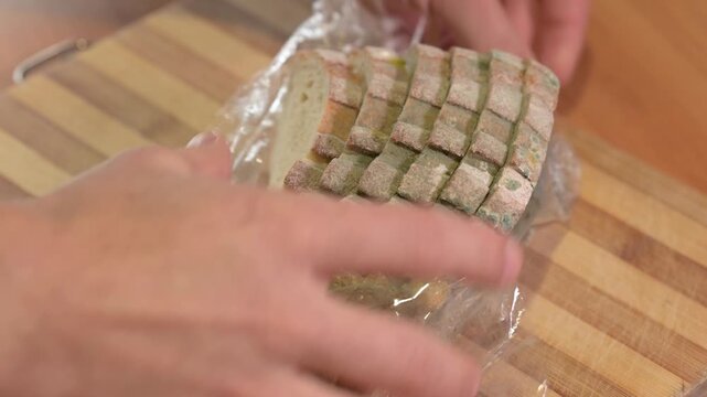 Man unpacking sliced white moldy bread from plastic on a board. Close-up shot of unpacking spoiled bread with green mold, symbolizing waste, health hazard, bacteria and food loss