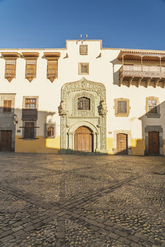View of the historic Casa de Colon with its ornate stone doorway, wooden balconies, and cobblestone square in Las Palmas de Gran Canaria, Canary Islands, Spain.