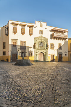 View of the historic Casa de Colon with its ornate stone doorway, traditional wooden balconies, and a fountain in the cobblestone square in Las Palmas de Gran Canaria, Canary Islands, Spain.