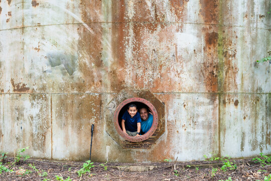 Father and son peering from a circular opening in a weathered, rusty industrial tank, enjoying an outdoor adventure