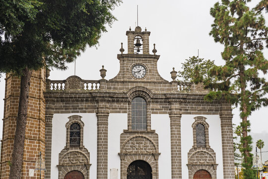 View of the Basilica of Our Lady of the Pine with its distinctive stone facade, arched entrances, and clock tower framed by tall pine trees in Teror, Canary Islands, Spain.