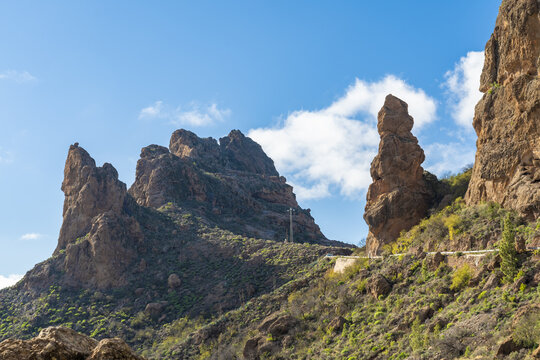 View of the impressive Roque Bentayga volcanic rock formation rising above the rugged landscape with sparse vegetation and a winding road under a blue sky in Tejeda, Canary Islands, Spain.
