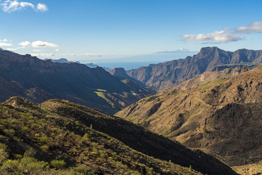 View of the rugged volcanic landscape surrounding Roque Bentayga with deep valleys and rocky slopes under a clear blue sky in Tejeda, Canary Islands, Spain.