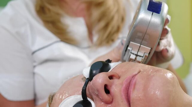 Close-up of female patient receiving IPL photorejuvenation. Cosmetologist applies silver device with blue accent to moist face for skin rejuvenation and anti-aging in clinic vertical 4k shot