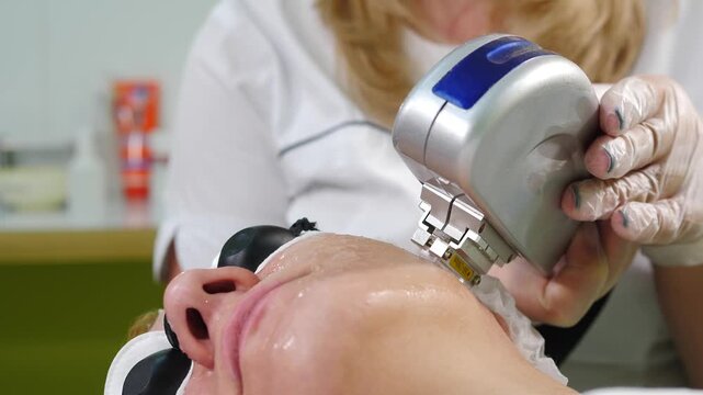 Cosmetologist gloved hand applying silver IPL device to female patient face. Skin covered with gel, protective eyewear on woman. Professional anti-aging photorejuvenation procedure vertical 4k shot
