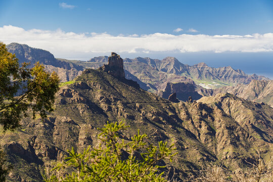 View of the rugged volcanic mountain landscape and the iconic Roque Nublo rock formation under a bright blue sky in Tejeda, Canary Islands, Spain.