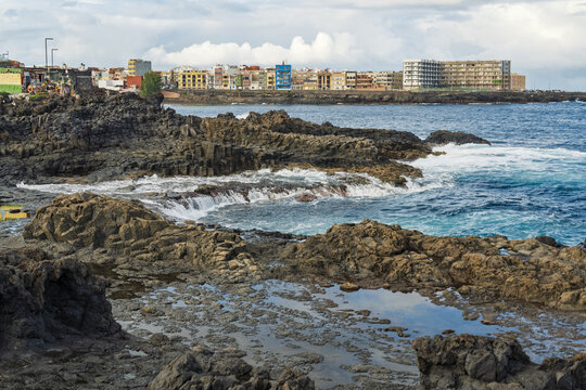 View of the volcanic rocky coastline and crashing ocean waves with colorful buildings in the background in La Garita, Canarias, Spain.