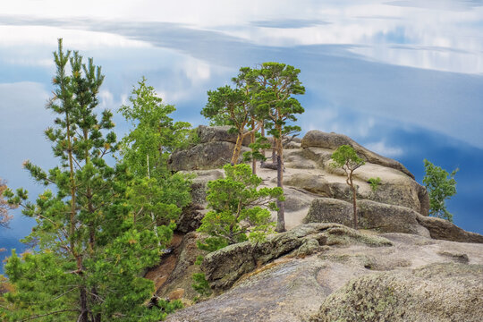 Green pine trees grows among stones on peak of mount Bolektay above Burabay Borovoe Lake, white cloud reflection on ripples blue water of lake. Burabay National Nature Park. Summer scene landscape.