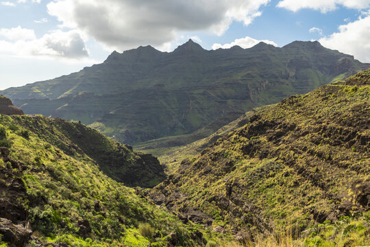 View of the rugged mountainous landscape of the Veneguera valley with rocky slopes, lush green vegetation, and a bright sky in Veneguera, Gran Canaria, Spain.