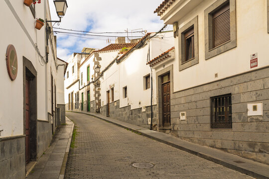 View of a quiet, narrow cobblestone street lined with traditional white-washed buildings featuring stone bases and wooden doors under a partly cloudy sky in Firgas, Canary Islands, Spain.