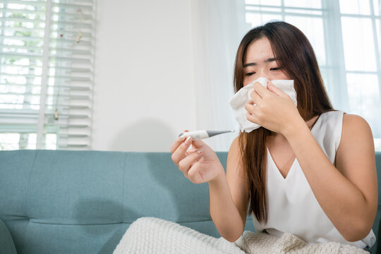 Sick Asian young woman sit under blanket on sofa she sneeze with digital thermometer