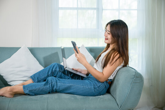 woman using smart mobile phone while sitting on sofa in living room at home with laptop computer