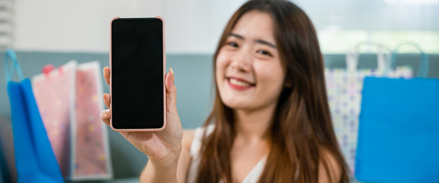 Excited Asian buying woman smiling showing mobile phone with many shopping bags at home