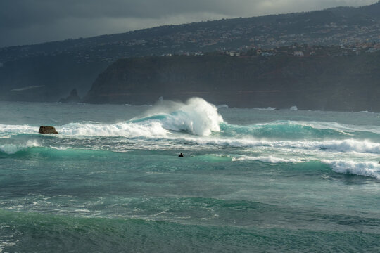 View of a surfer waiting for a wave in the turquoise ocean with dramatic crashing surf and rocky cliffs along the coastline in Puerto de la Cruz, Canary Islands, Spain.