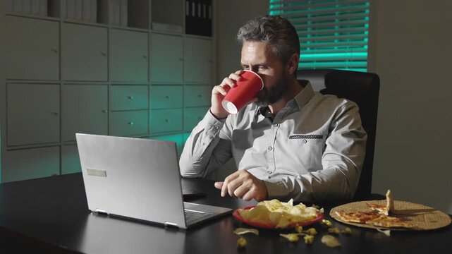 Office man eating and browsing web at night office. Businessman relaxing after work. Casual man lifestyle at night. Home office scene with late worker.