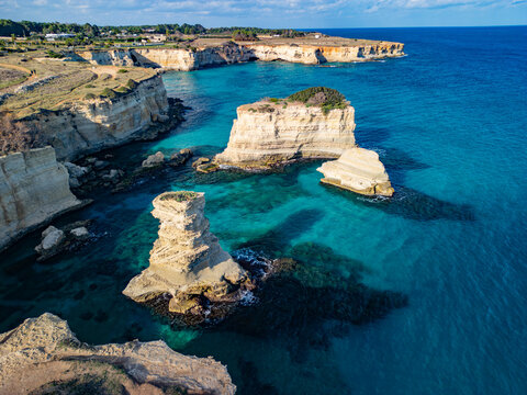 Aerial view of Beach Torre Sant'Andrea with dramatic limestone sea stacks rising from turquoise waters, rugged coastal cliffs, and clear blue sky in Melendugno, Puglia, Italy.