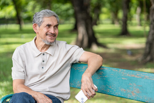 portrait elderly pensioner man spend leisure time relaxing in urban park,sitting on bench,holding mobile phone,smiling and looking in nature