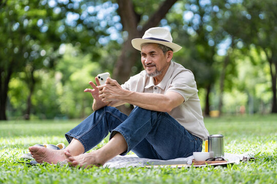 elderly pensioner man wearing hat sitting on lawn in urban park,surfing internet on smartphone,older adult spend leisure time relaxing in nature