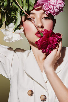 Fashion model with vivid makeup poses with pink and white flowers contrasting her pale cream jacket. Creative beauty concept with floral art elements and elegant styling.