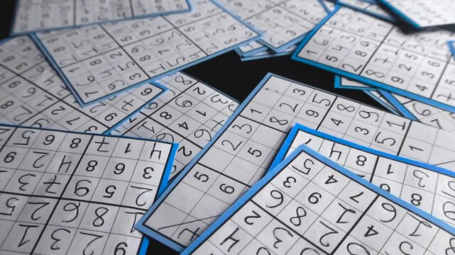 Numerous paper cutouts of solved sudoku puzzles with blue borders lying scattered on a black studio background, showcasing the complexity and solution of the popular number and logic game