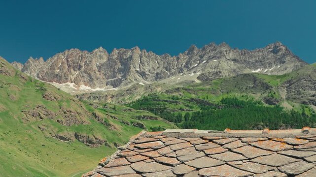 Static composition of a rugged Alpine mountain ridge with towering peaks rising above a rustic stone roof, creating a timeless mountain scene beneath a clear summer sky.