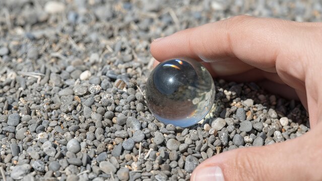 Hand Holding a Glass Sphere Over Pebbles on a Sunlit Surface