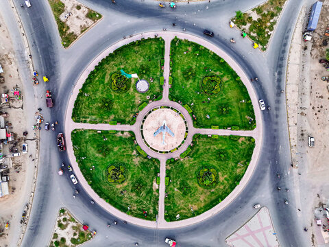 Aerial view of the Pakistan Chowk roundabout featuring a central fighter jet monument, green lawns, and a Pakistani flag, surrounded by traffic in Dera Ghazi Khan, Punjab, Pakistan.