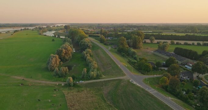 Drone flies above a Dutch dike road at sunset, revealing a tranquil canal, green farmland and soft pastel skies, capturing the beauty of the iconic Dutch polder landscape.