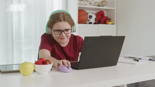 Excited Child uses a laptop for online learning. Amazed Child smiles while completing schoolwork at home. Funny child controls the computer mouse. Happy child studies at home.