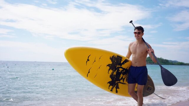 Caucasian young man walk along the beach carrying paddleboard and paddle. 