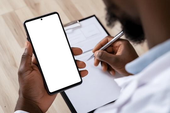 Close up doctor hands holding smartphone with blank white mock up screen writing on clipboard document at desk in clinic medical healthcare concept