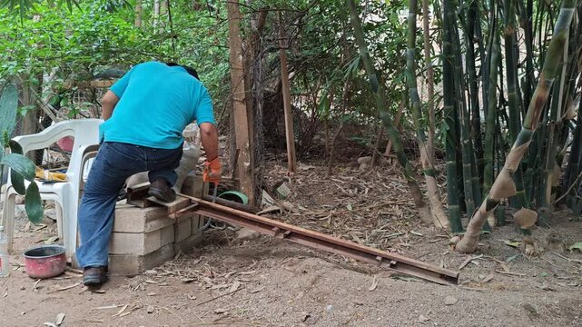 Young man working outdoors beside bamboo plants cutting a rusty iron bar