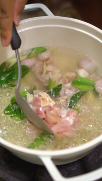 Cooking Thai spicy chicken tom yum soup. Boiling broth with lemongrass, galangal, shallots, and kaffir lime leaves, adding chicken pieces, sliced tomatoes, chili paste, and fresh basil leaves in a pot
