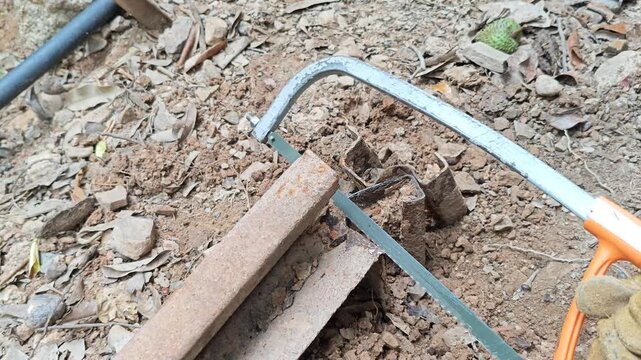 Manual hacksaw cutting through an oxidized iron pipe placed on dirt ground