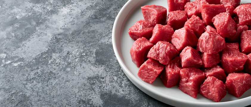 Top view flat lay of fresh raw diced beef meat cubes served on white plate over dark grey concrete background cooking recipe concept