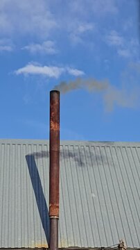 Thick black smoke from an old rusty chimney against a blue sky. Dark smoke rises from an old metal pipe above the roof of a building. Heating a rural bathhouse.