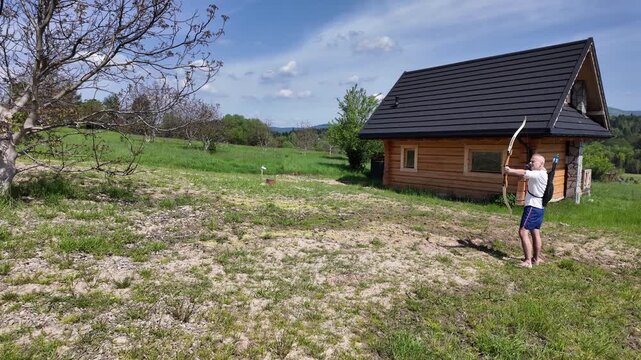 Archer practicing with a beginner-friendly recurve bow, retrieving arrows from a quiver between shots. Green grass, wooden cabin and bright summer landscape.