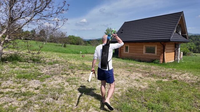 Archer practicing with a beginner-friendly recurve bow, reach for an arrow from a quiver between shots. Green grass, wooden cabin and bright summer landscape.