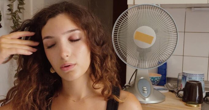 A woman wipes her forehead while feeling overheated indoors as an electric fan runs in the background during hot summer weather.