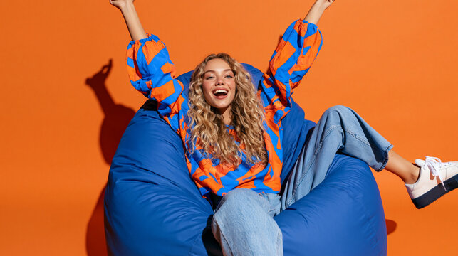 Joyful Exuberance: A young woman with a radiant smile and wild, curly blonde hair is captured mid-celebration, sitting on a large, plush beanbag chair against a vibrant orange backdrop.