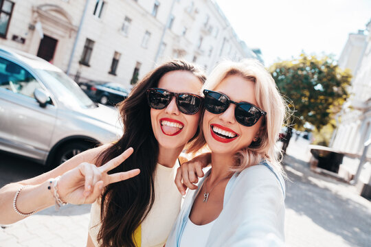 Two young beautiful smiling female. Sexy carefree women posing in the street. Positive models having fun, hugging in sunny day. With bright makeup and red lips, hold phone, take selfie photos