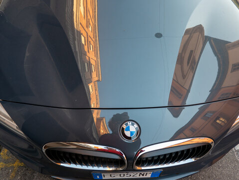 The BMW hood mirrors surrounding buildings, revealing the grille, brand emblem, and urban detail in a modern city
