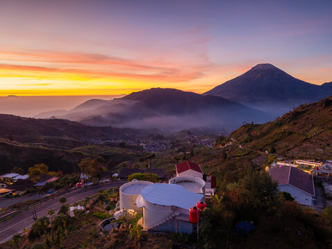 Aerial view of Sikapuk Hill and Villa Dieng featuring unique white circular buildings surrounded by rolling mountains and lush terraced hills under a vibrant sunset sky in Indonesia.