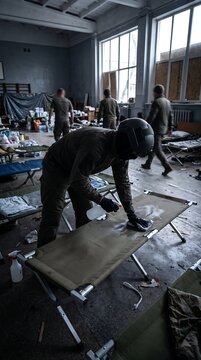Military personnel setting up folding cots in a temporary indoor field camp shelter