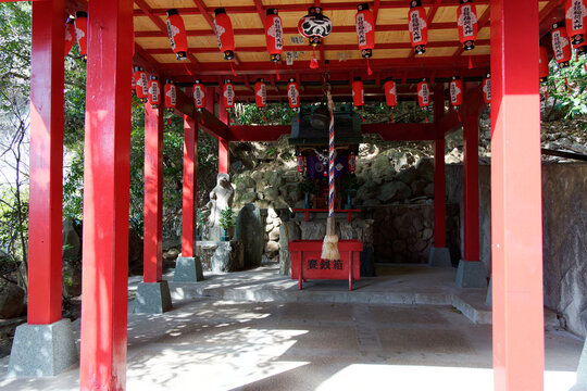 Traditional Japanese Shinto shrine worship hall with vibrant red pillars, hanging red paper lanterns, an offertory box, and a prayer bell rope under dappled sunlight.