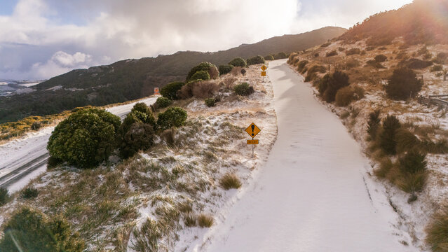Aerial view of Banks Peninsula road covered in fresh snow with a yellow warning sign and snow-dusted hills in the Canterbury Region, New Zealand.