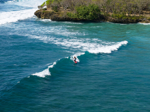 Aerial view of a surfer riding a wave at Nusa Dua Beach with turquoise water and a rocky coastline in Kecamatan Kuta Selatan, Bali, Indonesia.