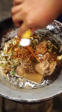 Reheating braised chicken soup in a wok. Pouring brown broth with braised chicken, bean sprouts, and radish, seasoning with chili flakes and sauce, adding thin rice vermicelli noodles, and boiling unt