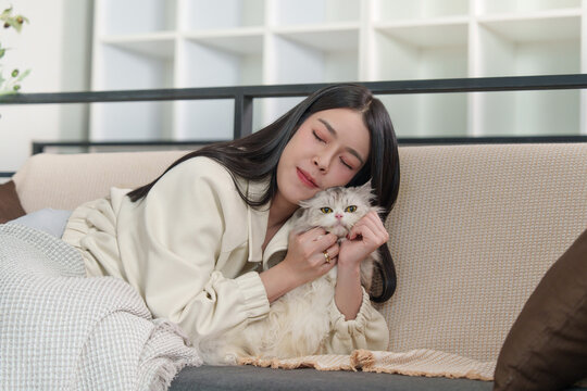 Asian woman cuddling fluffy cat with eyes closed on living room sofa