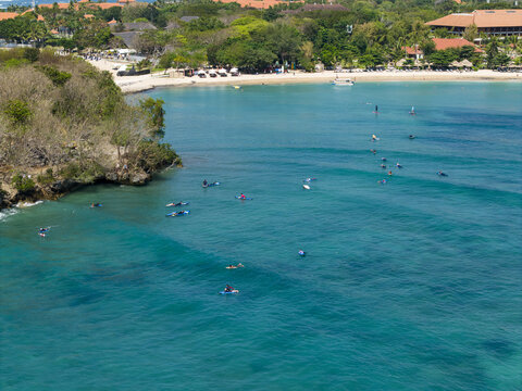 Aerial view of surfers on surfboards in the turquoise waters of Nusa Dua Beach with a sandy coastline and resort architecture in Kecamatan Kuta Selatan, Bali, Indonesia.
